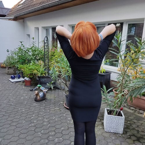 Person in schwarzem Kleid streckt sich im Garten, rotes Haar, Pflanzentöpfe umgeben sie.