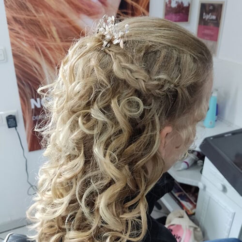 Person mit lockigem blondem Haar, verziert mit einem Perlen-Haaraccessoire, lächelt leicht.